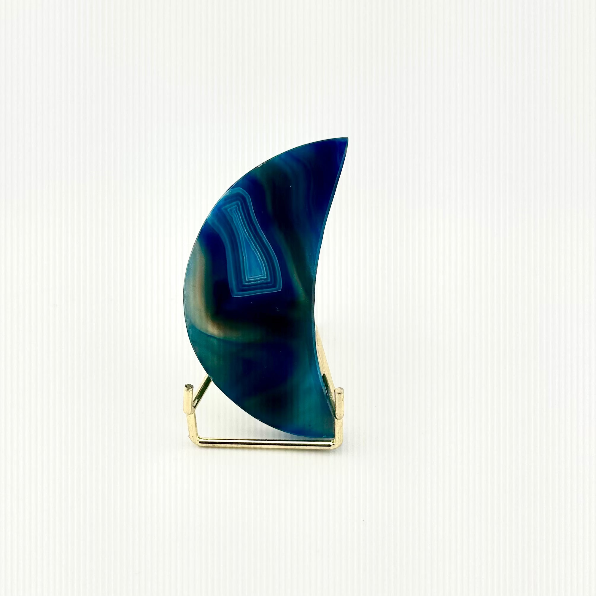 A crescent-shaped agate stone ornament in shades of deep blue and teal, displayed on a small gold metal stand against a clean white background.